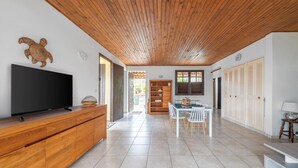 A spacious living area with a wooden ceiling, a flat-screen TV on a wooden cabinet, a dining table with chairs, and a bookshelf. - Villa Zenaida, a villa near the sea (Sainte-Luce)