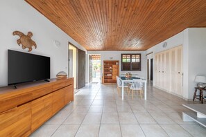 Uma sala de estar espaçosa com teto de madeira, uma televisão de tela plana sobre um móvel de madeira, uma mesa de jantar com cadeiras e uma estante de livros.