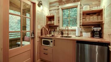 A compact kitchen with wooden cabinetry, a built-in microwave, a stainless steel dishwasher, and a window with a view of trees.
