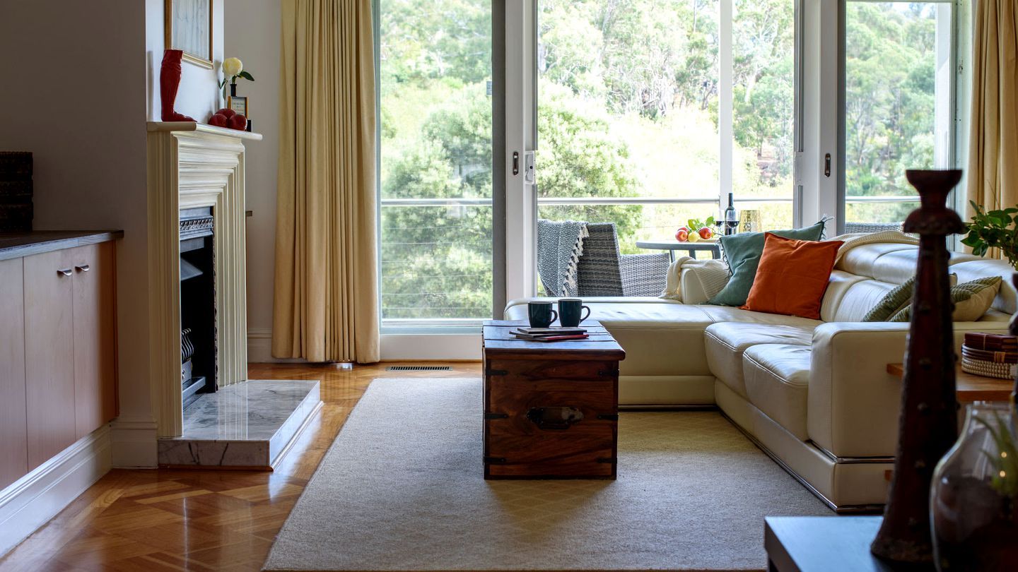 A living room with a sofa, a coffee table, and a fireplace. There is a view of trees through large windows.