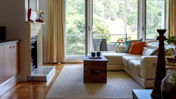 Een woonkamer met een bank, een salontafel en een open haard. Er is uitzicht op bomen door grote ramen.