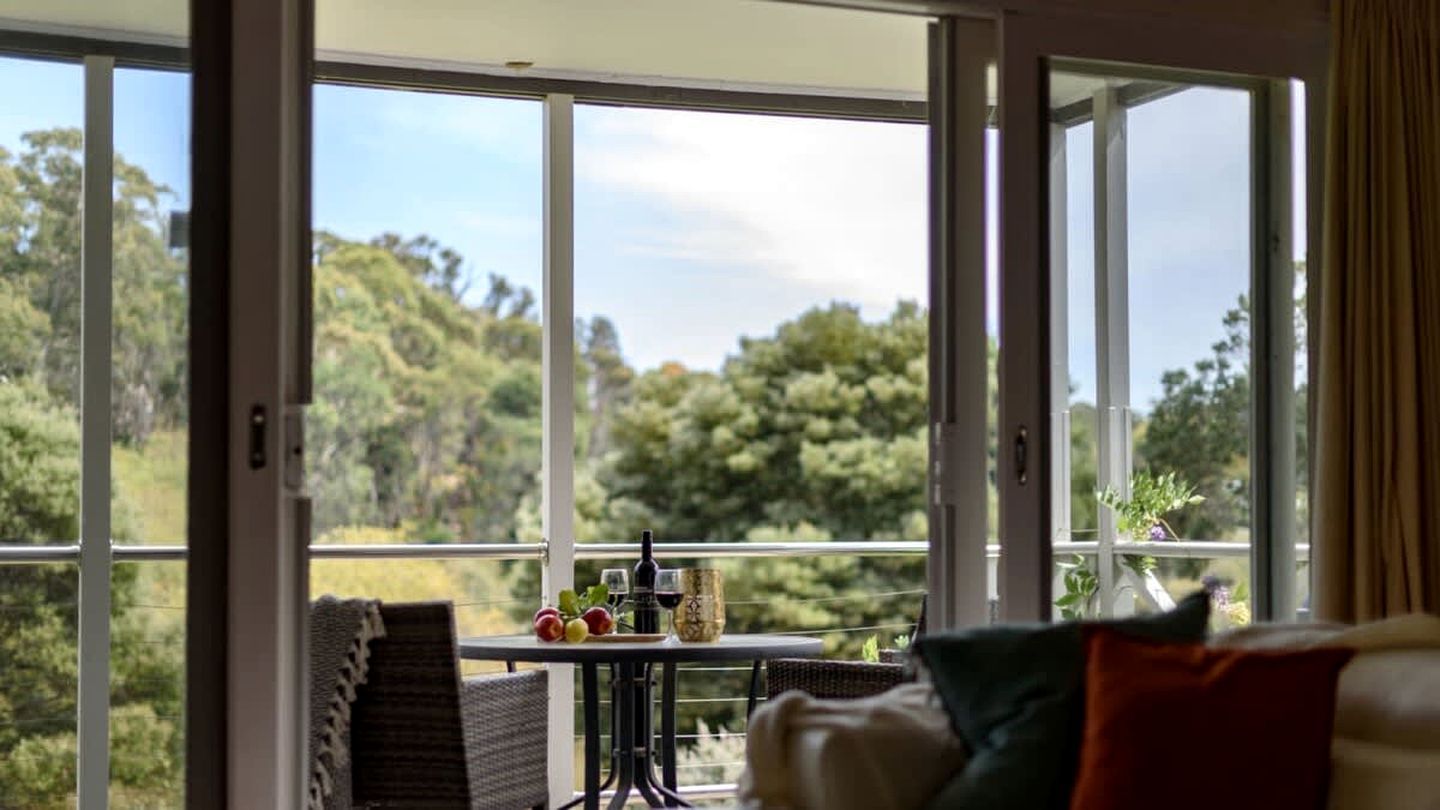 A room with a large window, a table with fruits and a bottle, and a sofa with cushions.