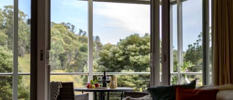 A room with a large window, a table with fruits and a bottle, and a sofa with cushions.