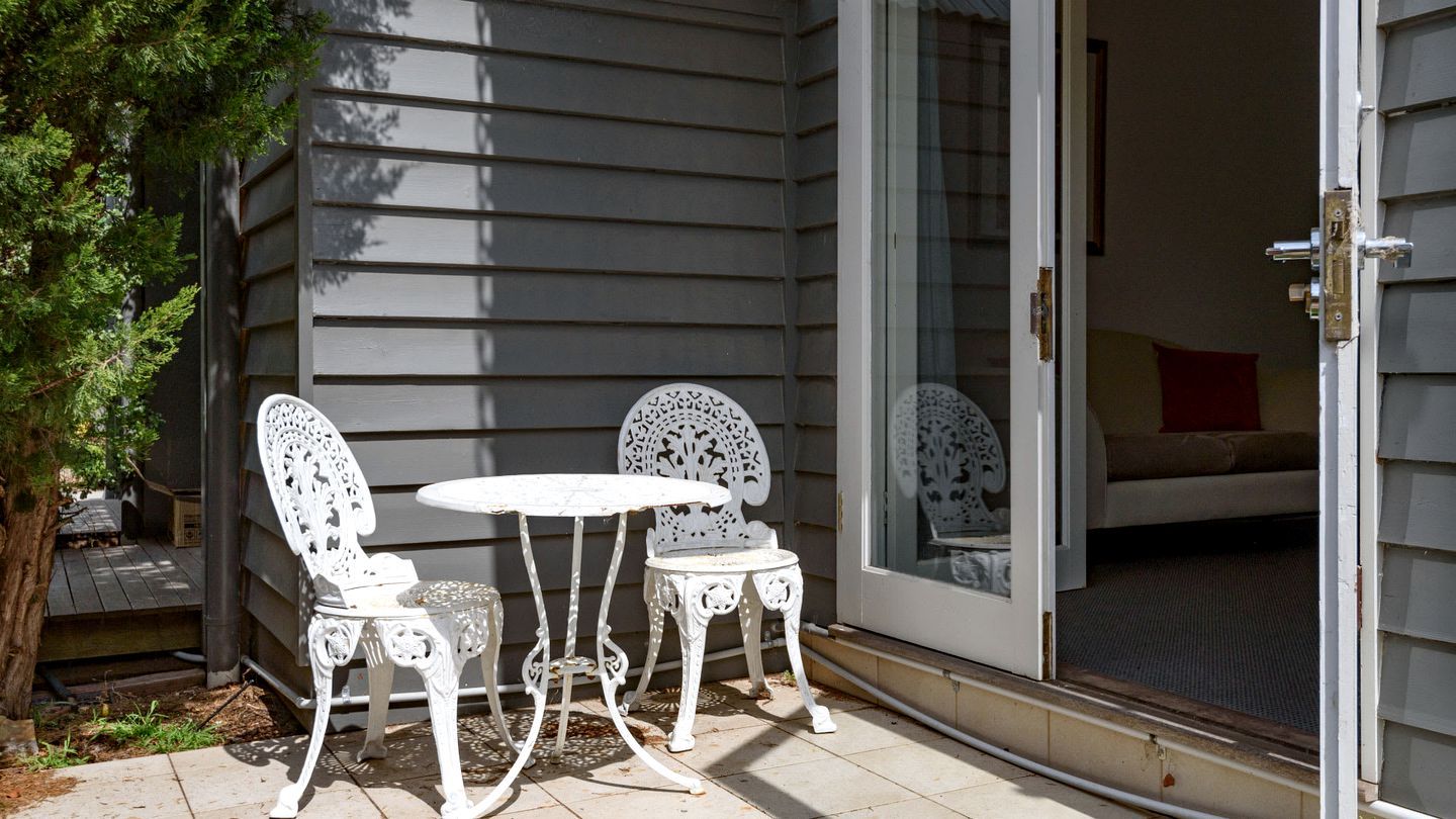 A patio with two white chairs and a table, a sliding glass door, and a small tree.