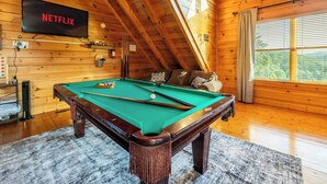 Une table de billard dans une pièce aux murs en bois, un téléviseur sur lequel on peut lire le mot « NETFLIX », et un panneau indiquant « SALLE D’ACCÈS ».