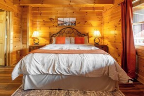 A wooden cabin bedroom with a bed, two bedside lamps, a window with curtains, and a painting on the wall. - Mtn Views! Hot tub! 2 Pools! Mini Golf! Family fun (Sevierville)