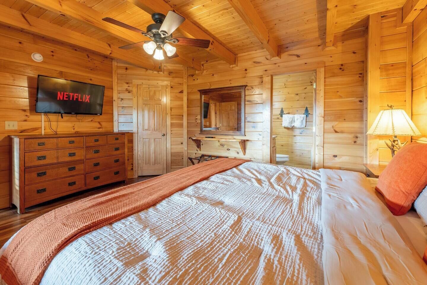 Una habitación de cabaña de madera con una cama, un tocador, un televisor y un ventilador de techo.