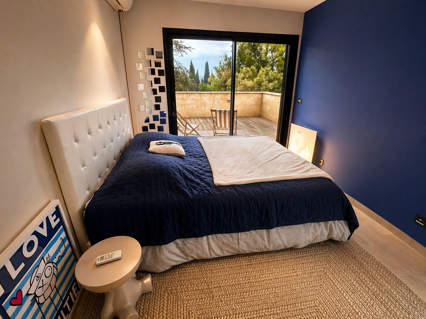 Une chambre à coucher avec un lit, une petite table, un élément de décoration murale et une vue sur l’extérieur.