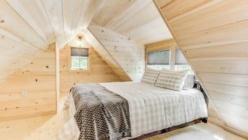 A bedroom with a sloped ceiling, wooden walls, a bed with a checkered blanket, and two windows with blinds.