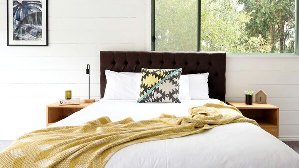 A bed with a tufted headboard, white bedding, a patterned pillow, a yellow blanket, and a bedside table with a lamp and a small plant.
