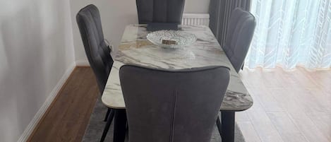 A dining area with a marble table, grey upholstered chairs, a grey rug, and a window with grey curtains.