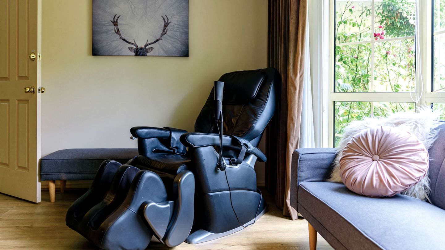 A massage chair in a room with a window, a sofa with a cushion, and a door.