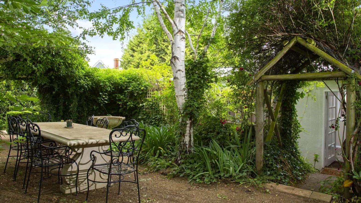 An outdoor garden with a table and chairs, surrounded by lush greenery and a wooden pergola.