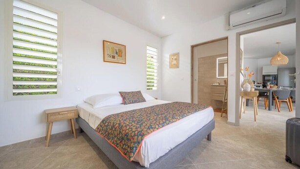 A bedroom with a bed, a bedside table, a window with blinds, and a door leading to a kitchen area. - Creole Villas (10 people)•Swimming Pools•Beach within walking distance (Sainte-Luce)