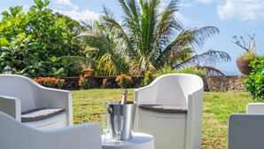 Ein Sitzbereich im Freien mit weißen Stühlen und einem Tisch, einer Champagnerflasche und Gläsern, vor einer Kulisse aus üppigem Grün und einer Steinmauer.