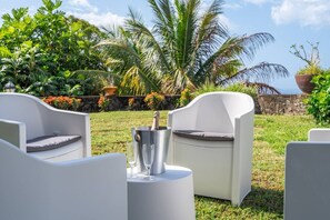 Ein Sitzbereich im Freien mit weißen Stühlen und einem Tisch, einer Champagnerflasche und Gläsern, vor einer Kulisse aus üppigem Grün und einer Steinmauer.