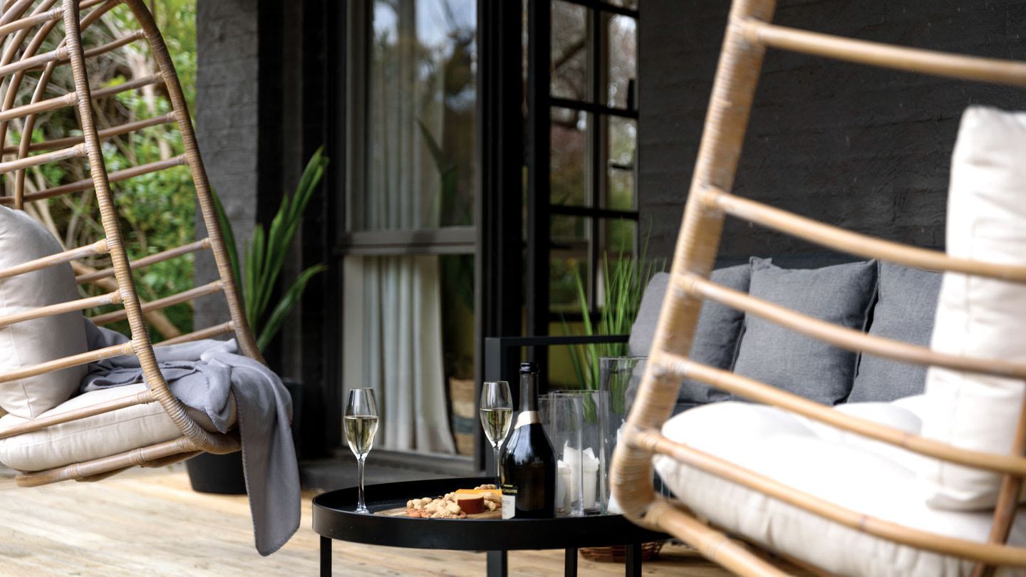 Una silla de mimbre con cojines, una mesita redonda con una botella y vasos, y una ventana con vistas a la vegetación.