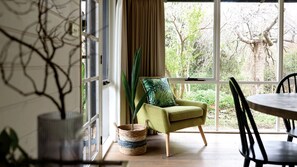 Ein Zimmer mit großem Fenster, einem grünen Sessel, einem Korbwägelchen und einem Esstisch mit Stühlen.