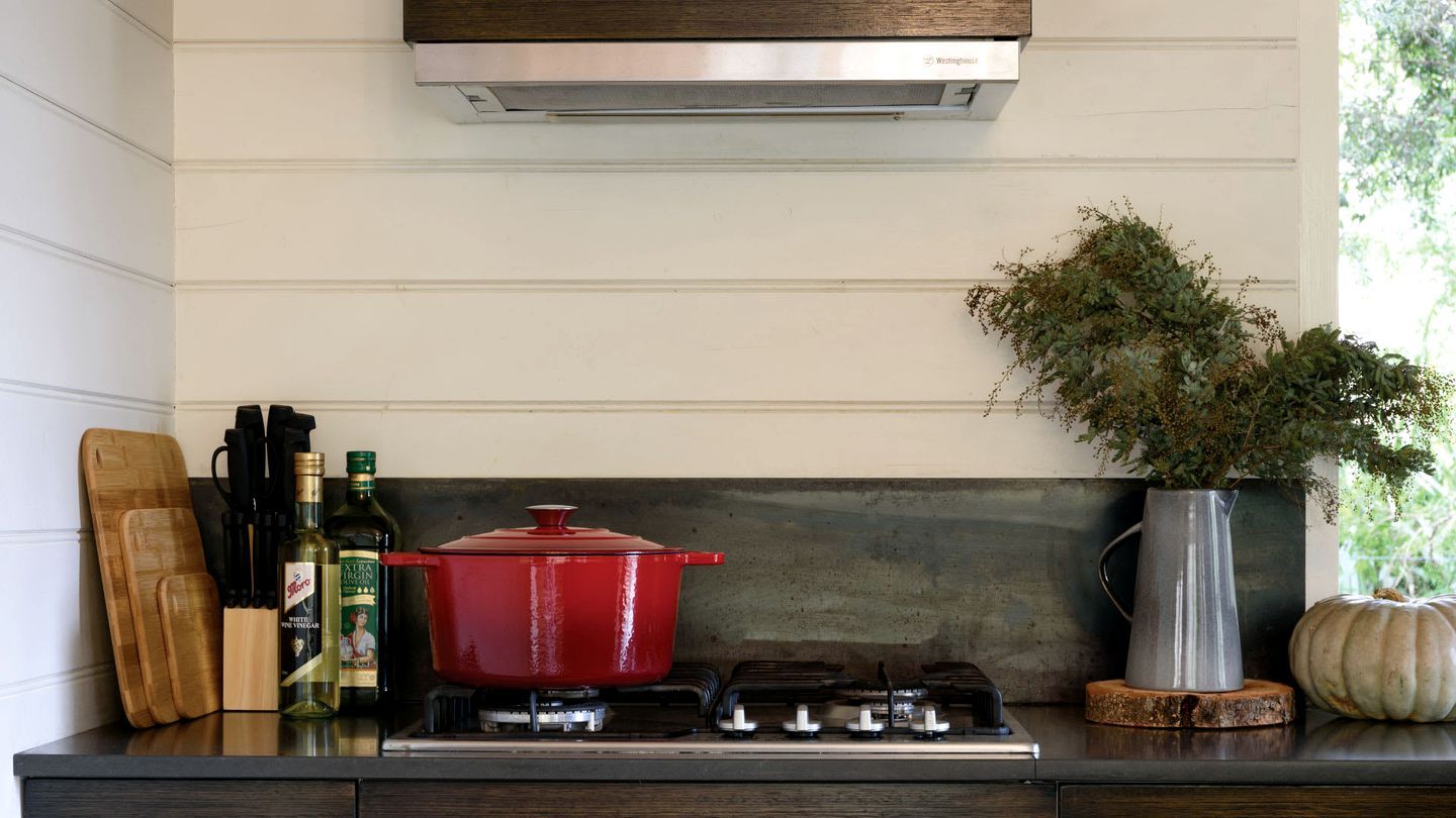Une cuisine avec une casserole rouge sur la cuisinière, une bouteille d’huile d’olive et une plante en pot.
