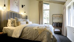 Ein Schlafzimmer mit Bett, Nachttischen, einer Wandleuchte, einem Fenster mit Vorhängen und Blick ins Grüne.