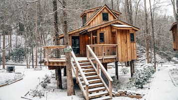 Une cabane en bois dotée d’une terrasse et d’un escalier, entourée d’une forêt enneigée.