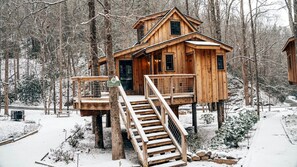 A wooden cabin with a deck and stairs, surrounded by a snowy forest.