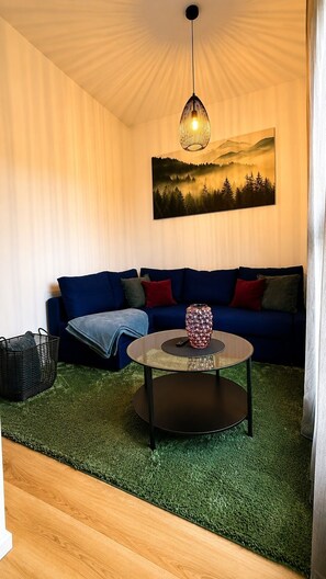 Una habitación con un sofá azul, una mesa redonda de centro con un jarrón decorativo, una lámpara colgante y un cuadro de un paisaje forestal.