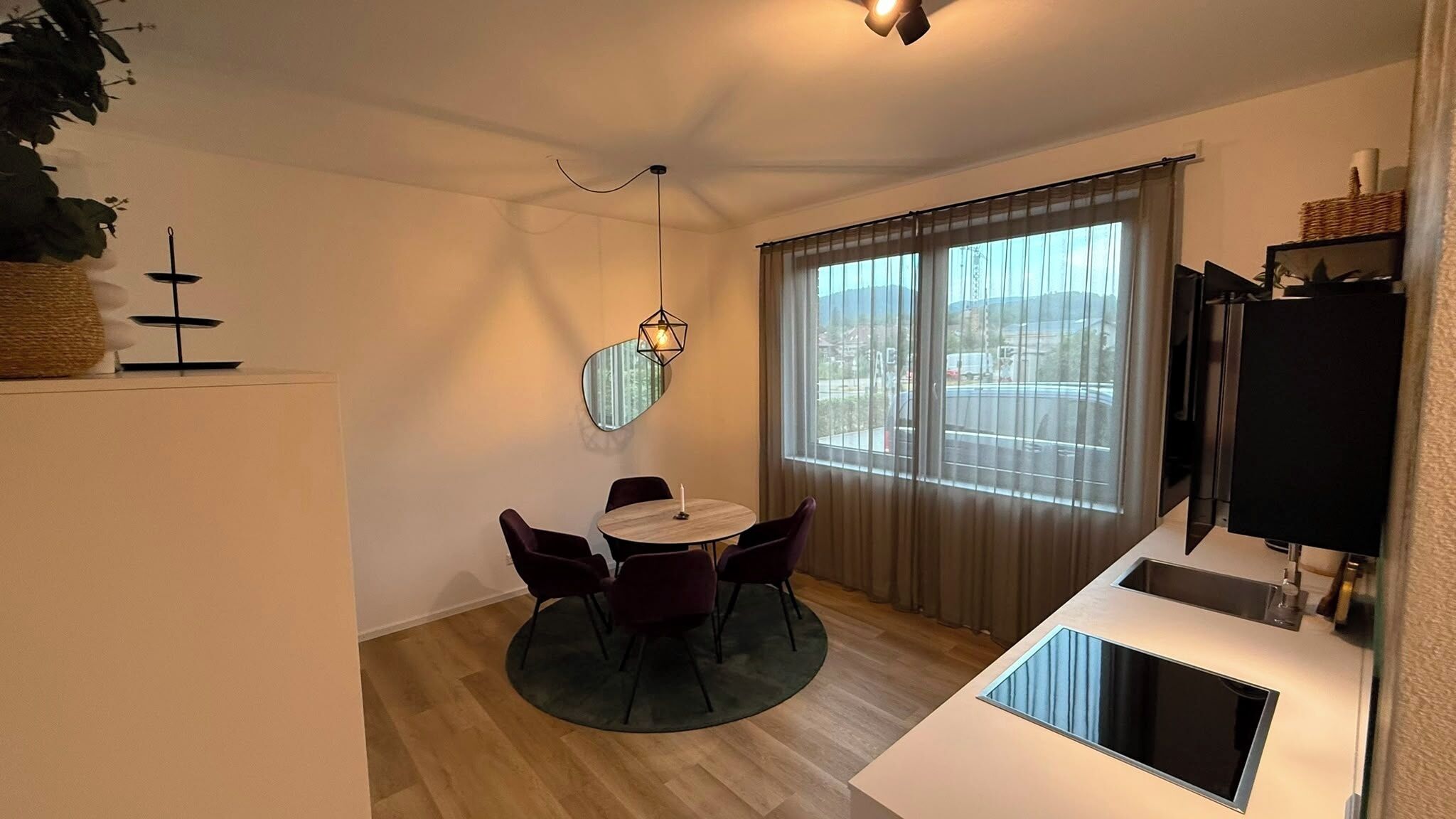 A compact living space with a dining area, a small kitchen, and a large window with curtains.