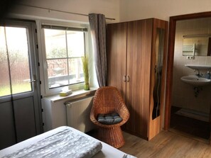Ein Schlafzimmer mit einem Bett, einem Korbstuhl, einem Kleiderschrank und einem Badezimmer mit Waschbecken und Spiegel.