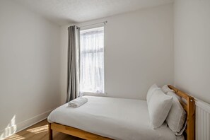 Ein kleines Zimmer mit einem Einzelbett, einem Holzkopfstück, einem Fenster mit durchsichtigen Vorhängen und einem Heizkörper an der Wand.