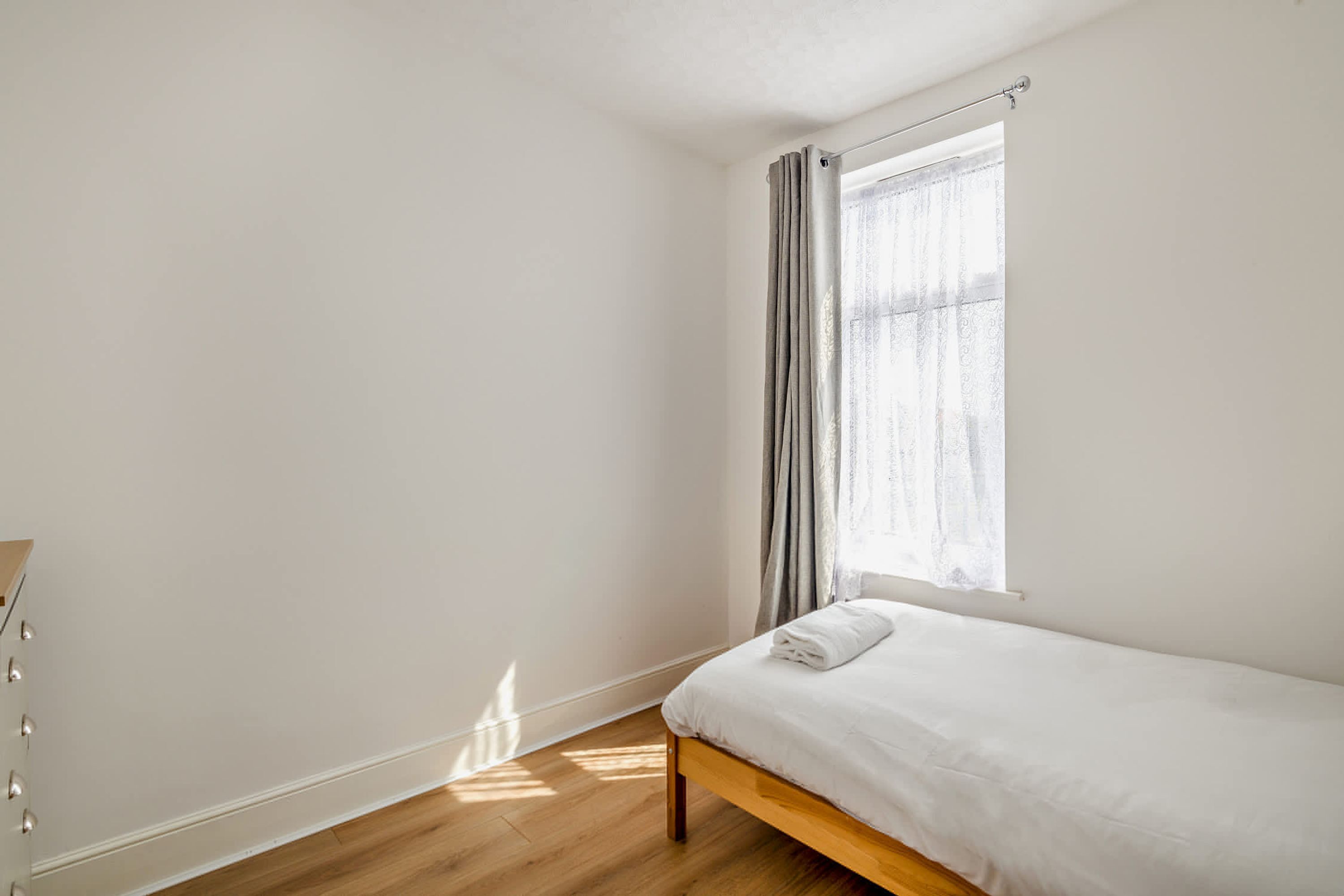Ein schlicht eingerichtetes Schlafzimmer mit einem Bett, einem Fenster mit Vorhängen und Holzboden.