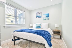 A bedroom with a bed, a window with blinds, two wall-mounted lamps, and two framed pictures. - Seaside Retreat: Salty Shore Getaway (Wildwood)