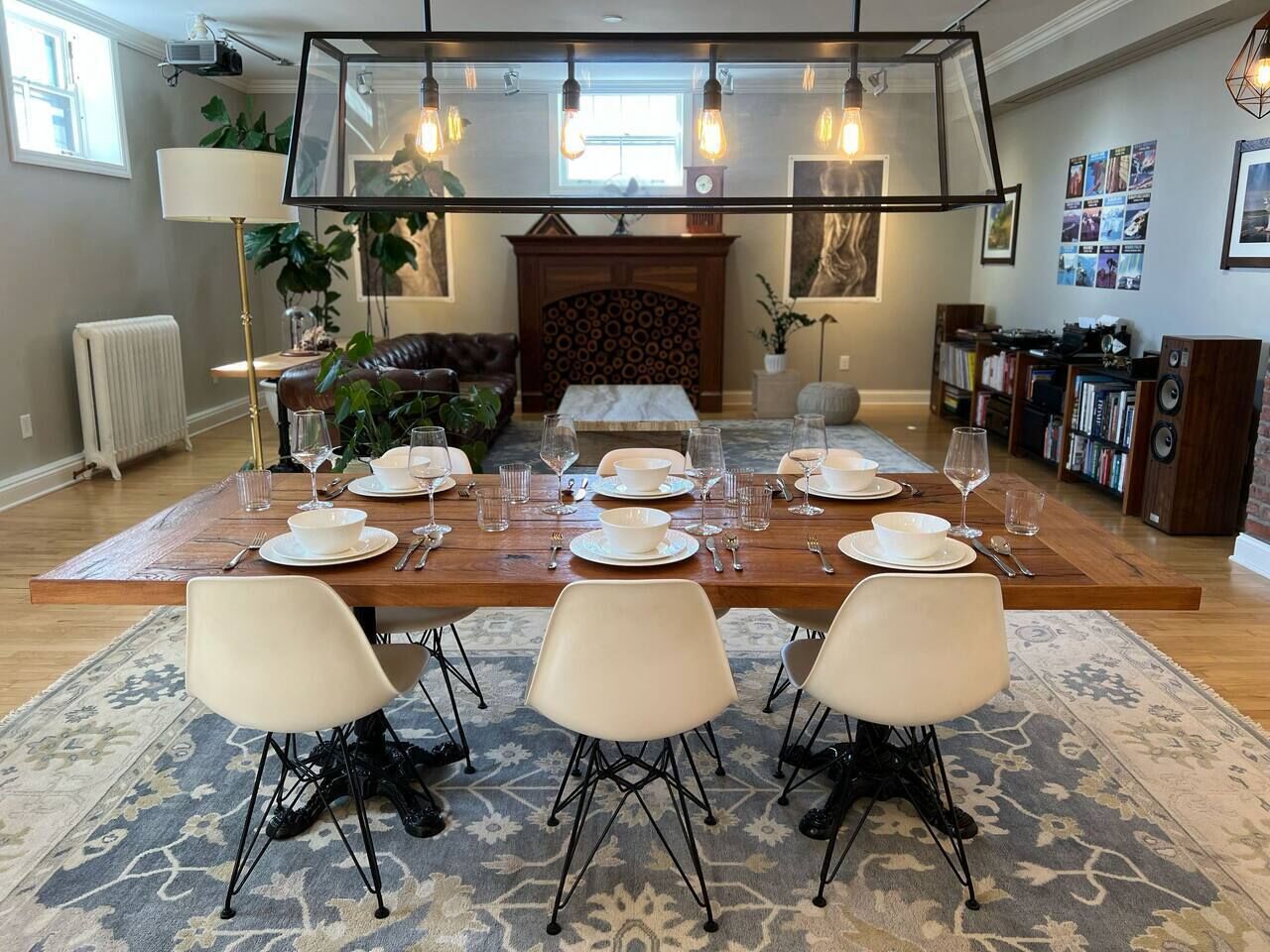 Un comedor con una larga mesa de madera dispuesta para cuatro, rodeada de sillas modernas, una chimenea y una estantería.