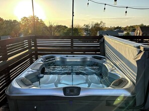 Un bain à remous doté d’un panneau de commande, entouré d’une terrasse en bois, avec vue sur des bâtiments et des arbres en arrière-plan.
