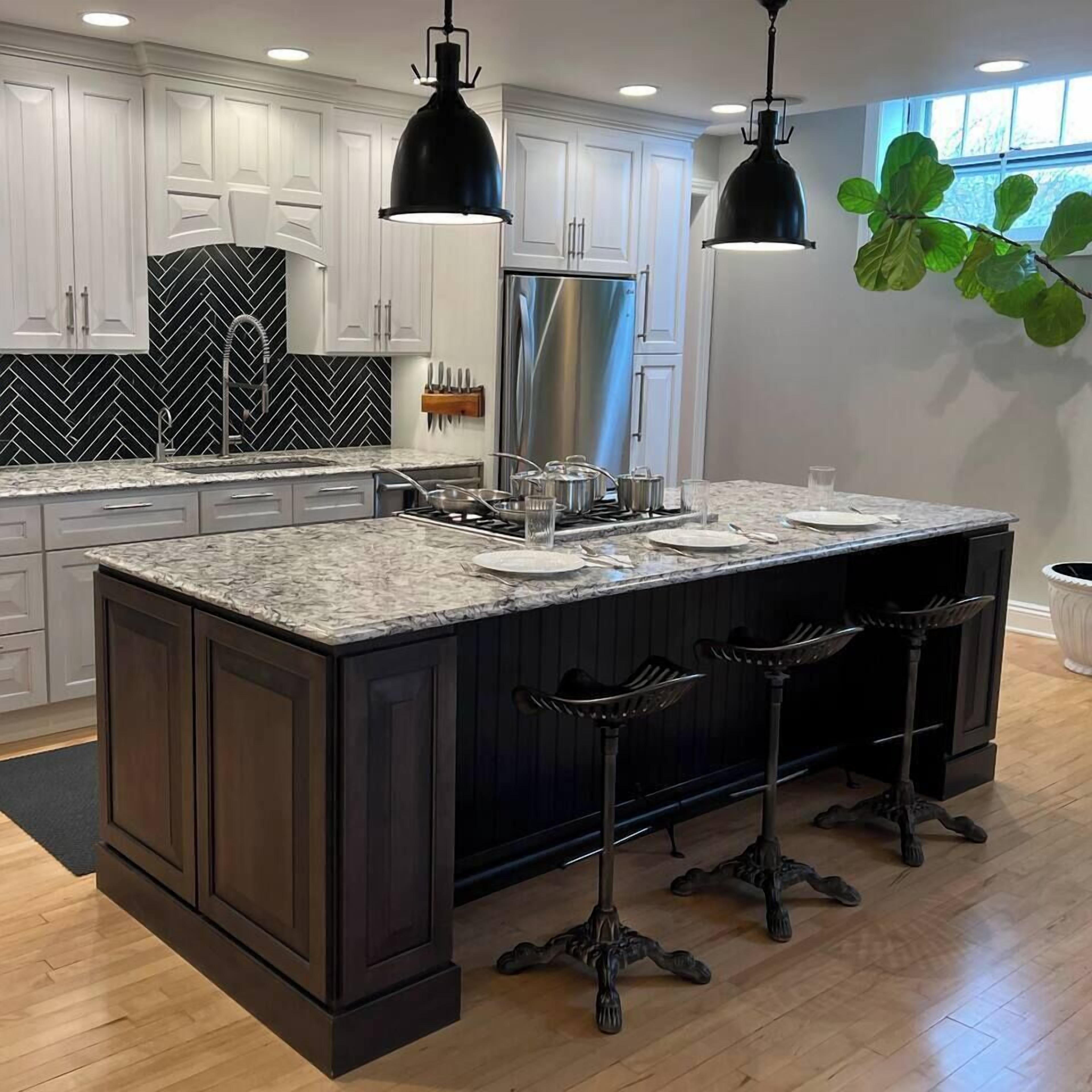 Una cocina con isla central, muebles oscuros, armarios superiores blancos, salpicadero de listones negros y electrodomésticos de acero inoxidable, con iluminación colgante.