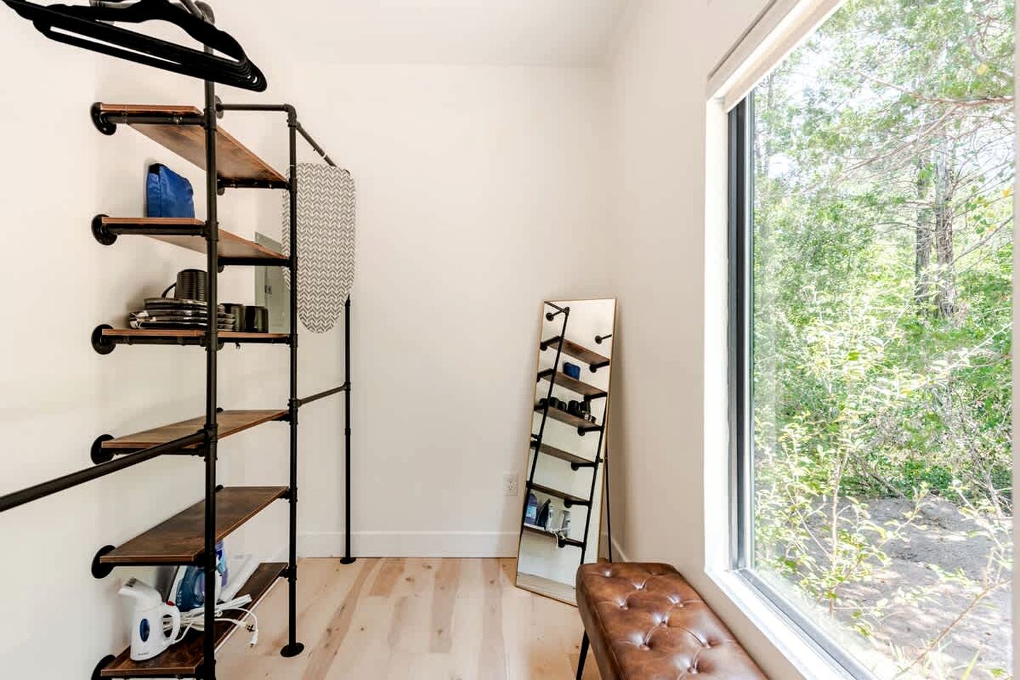 Ein Zimmer mit Holzboden, einem schwarzen Metallregal mit mehreren Ablagen, einer Bank und einem Fenster mit Blick auf Bäume.