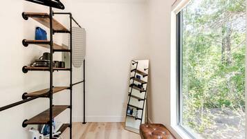 Ein Zimmer mit Holzboden, einem schwarzen Metallregal mit mehreren Ablagen, einer Bank und einem Fenster mit Blick auf Bäume.