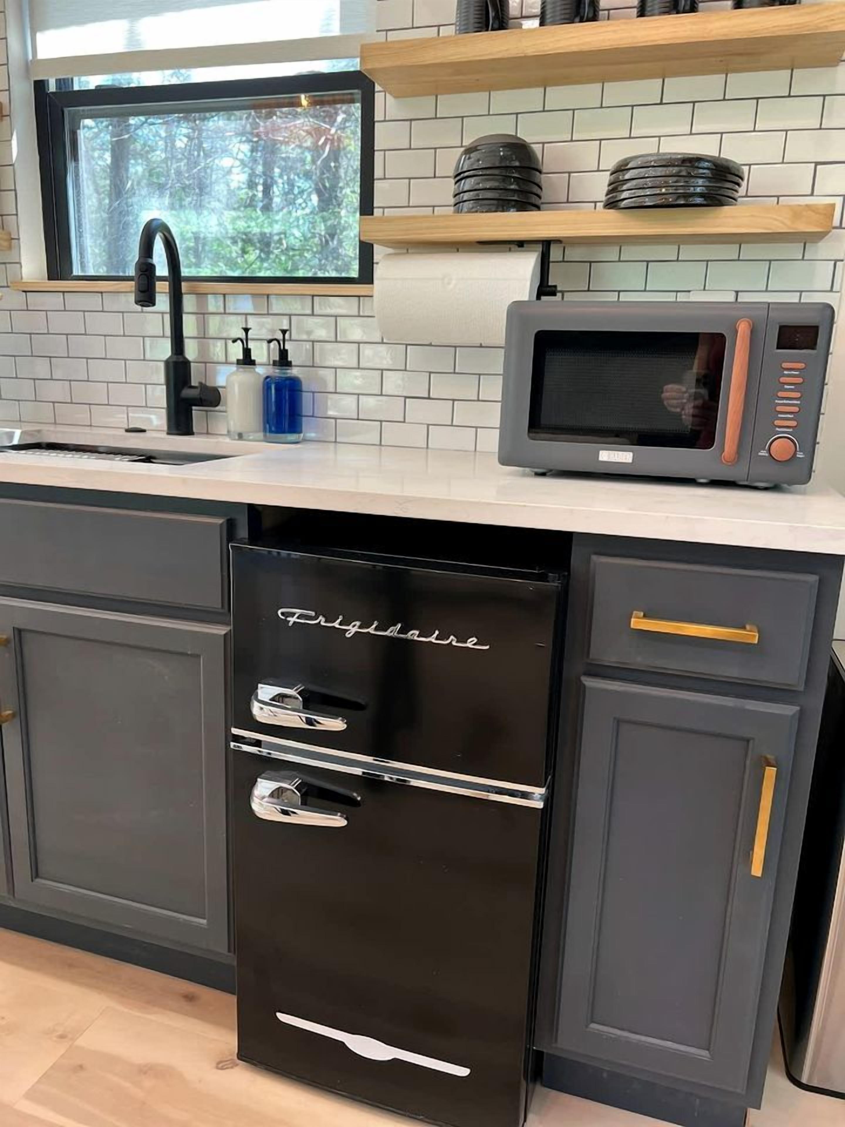 A kitchen with a black refrigerator, a microwave, a sink, and a window with a view of trees.