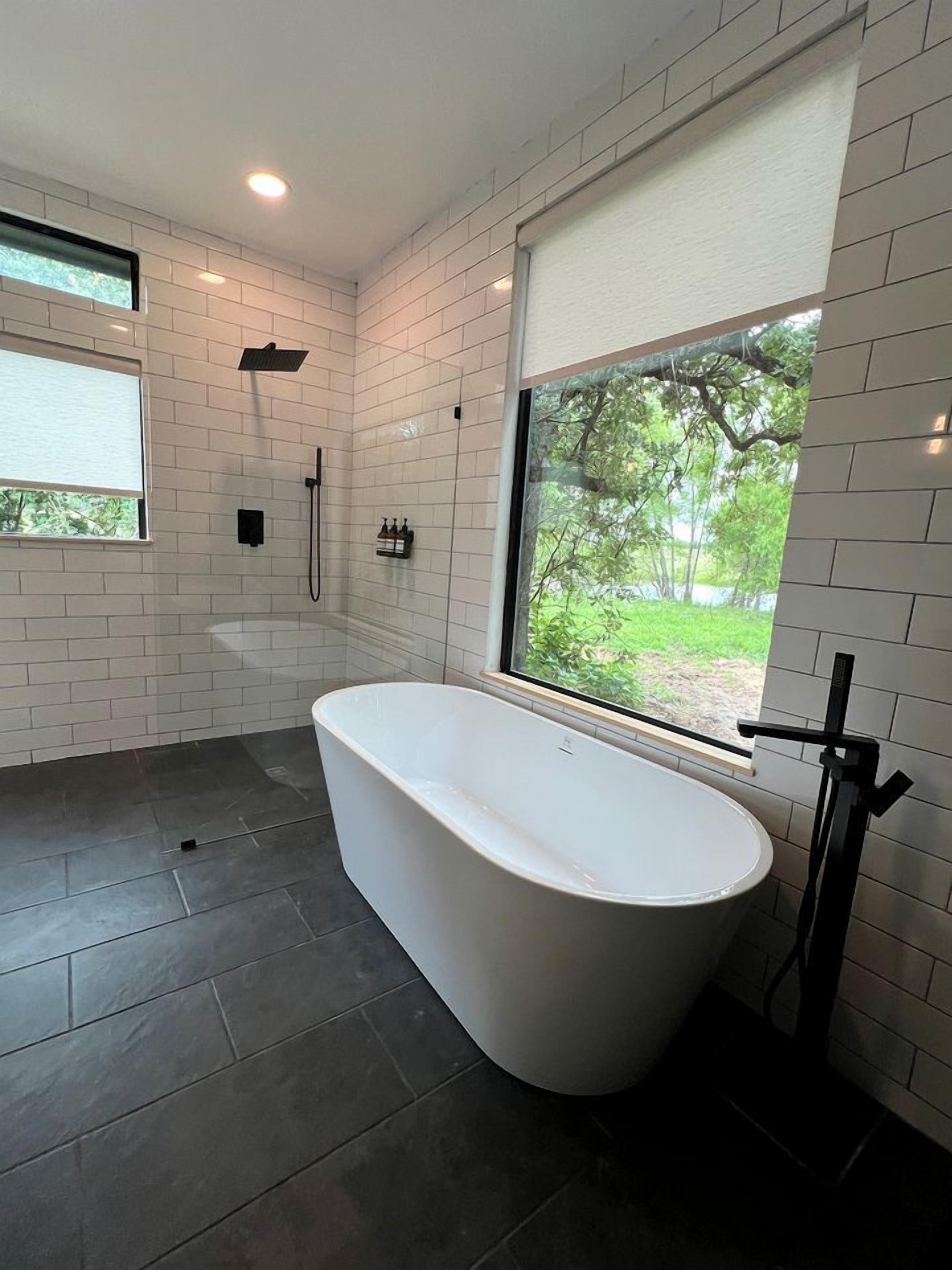 A modern bathroom with a freestanding bathtub, a shower, and a window providing a view of greenery outside.