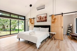 A modern bedroom with a large bed, a small round table, and a view of greenery outside.