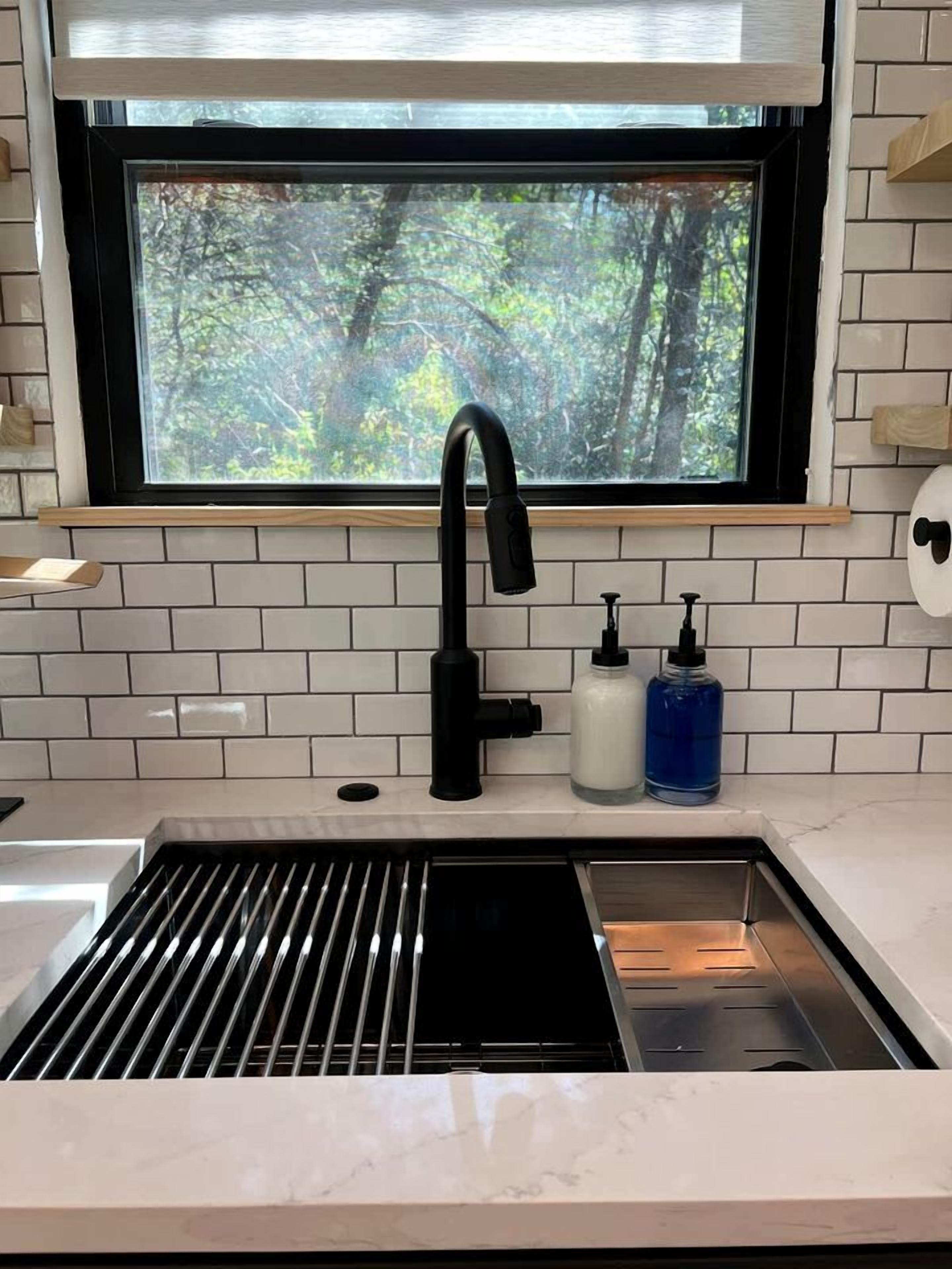 A kitchen with a black faucet, white tiles, and a window with a view of trees.