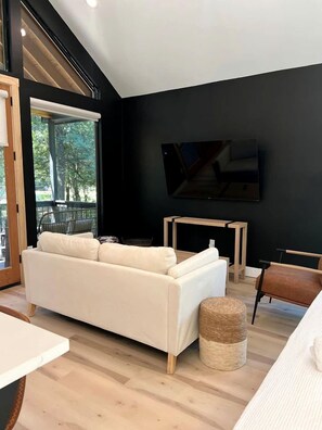 A modern living room with a white sofa, a small round ottoman, a wooden coffee table, and a flat-screen TV mounted on the wall.