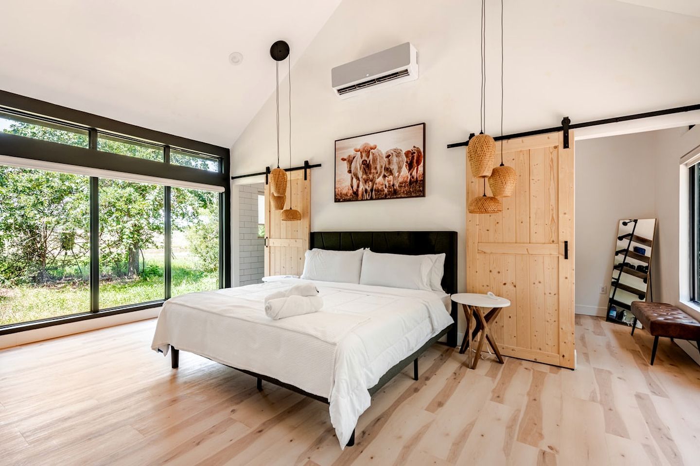 A modern bedroom with a large bed, a small side table, a wooden wardrobe, and a large window offering a view of greenery.
