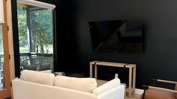 A modern living room with a white sofa, a small round ottoman, a wooden coffee table, and a flat-screen TV mounted on the wall.