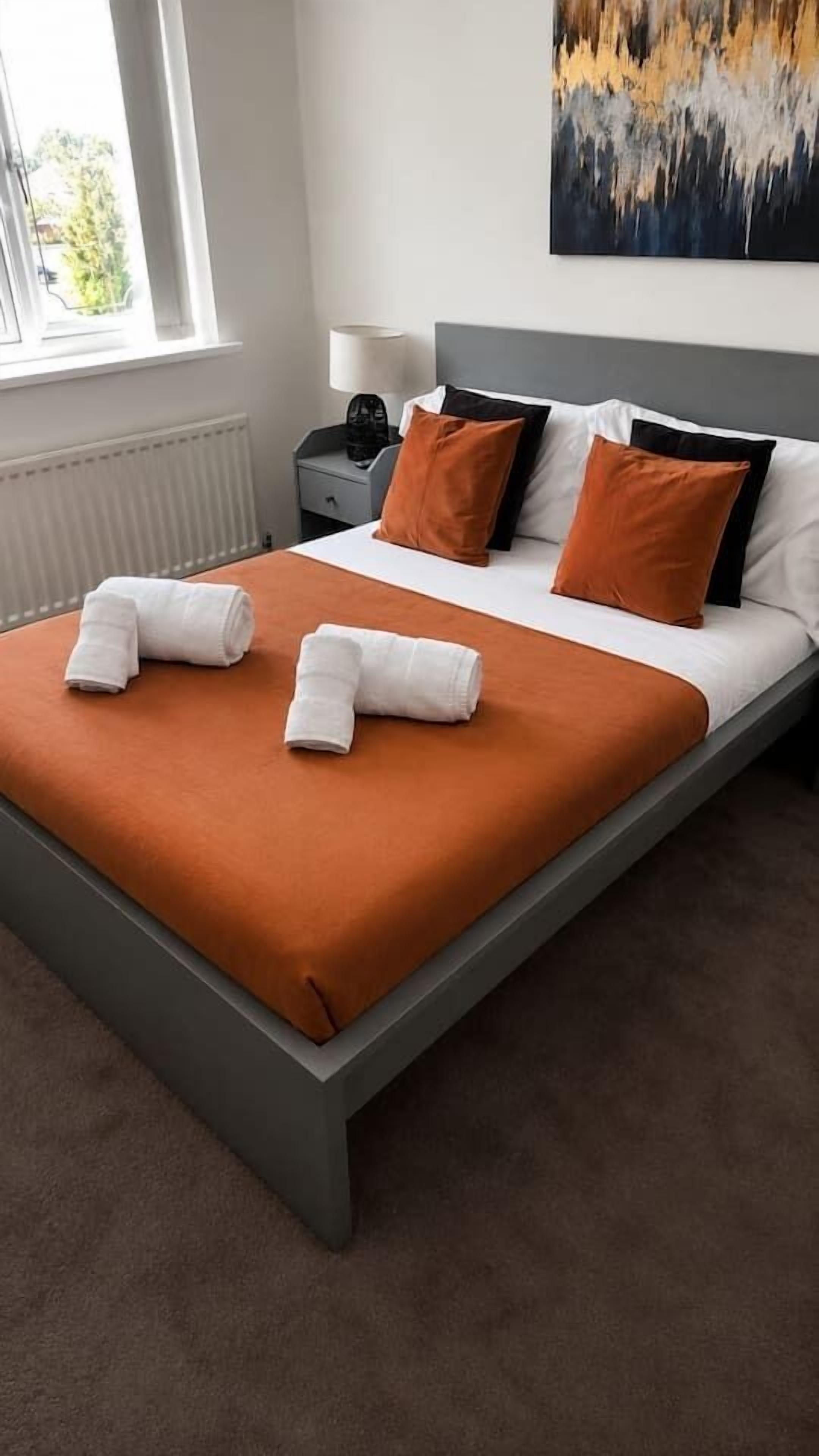 A neatly made bed with white and orange bedding, a gray headboard, and a painting on the wall.