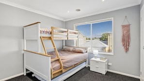 Ein Etagenbett mit einer Leiter, ein Nachttisch und ein Wandbehang aus Makramee in einem Zimmer mit Fenster und Blick auf ein Haus.