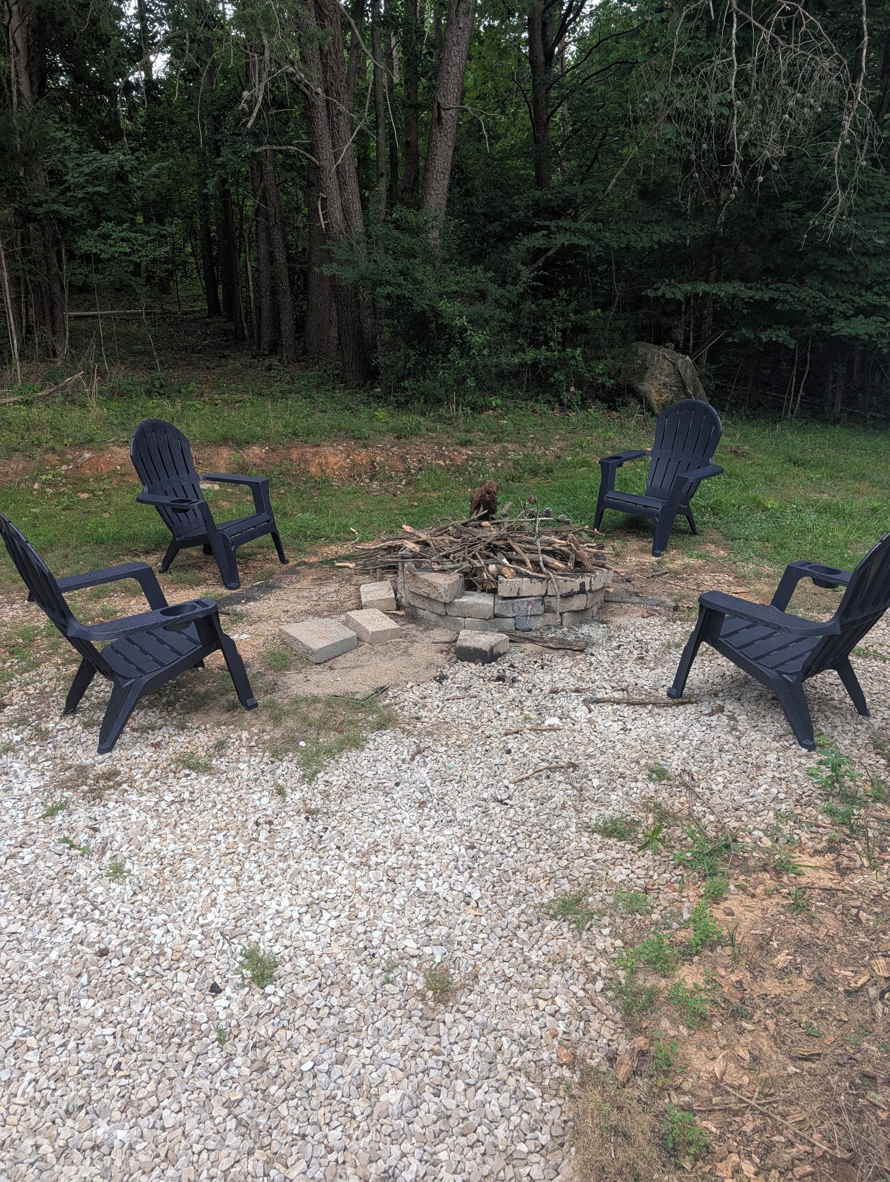 Et bålsted omgivet af fire Adirondack-stole i et skovområde.