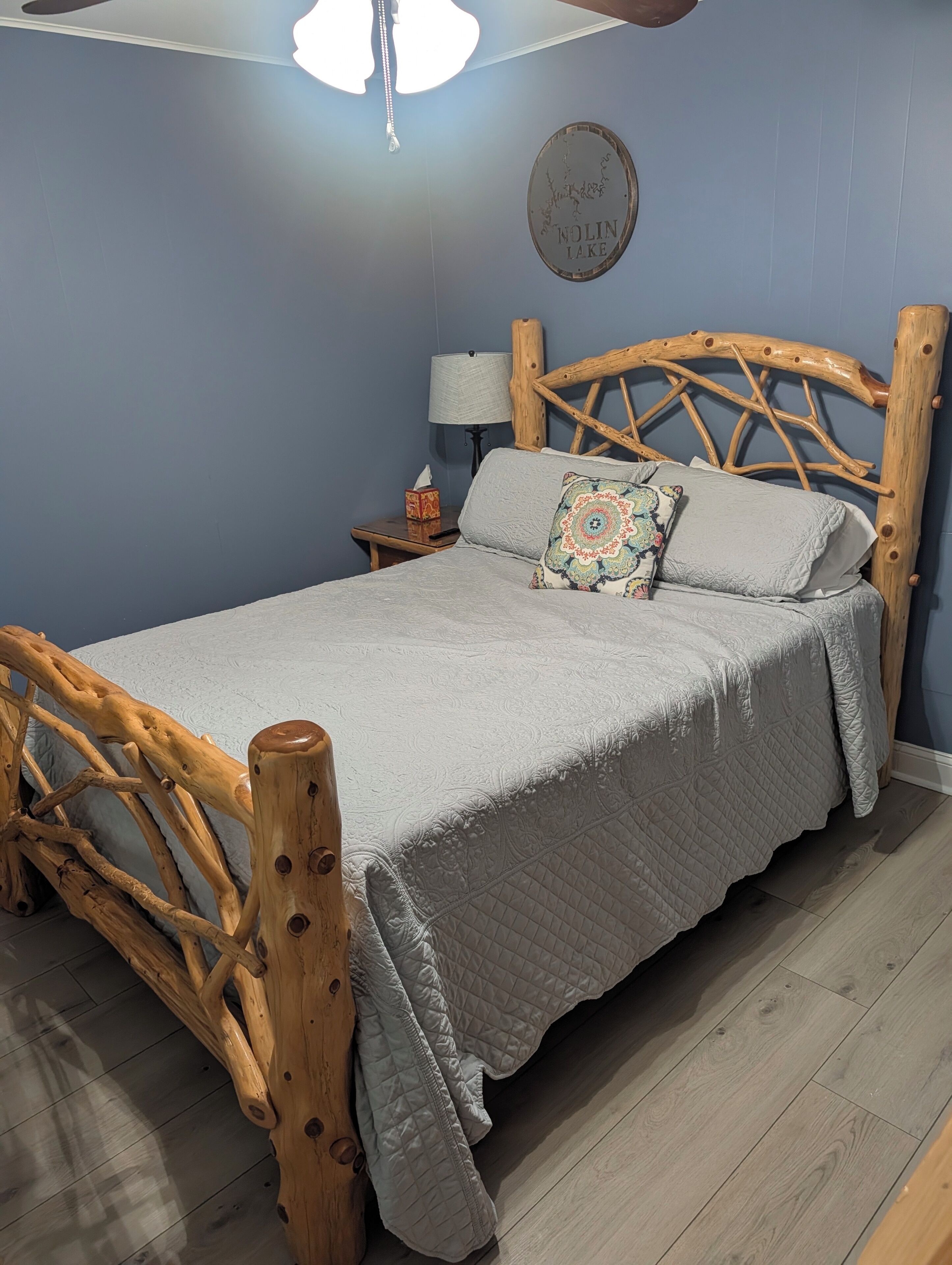 Un dormitorio con cabecera de cama de madera, colcha acolchada gris, almohada decorativa, mesita de noche con lámpara y un cartel en la pared.