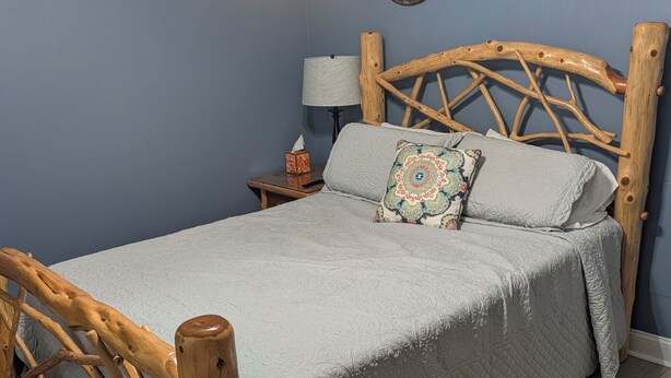 A bedroom with a wooden bed frame, a grey quilted bedspread, a decorative pillow, a bedside table with a lamp, and a wall-mounted sign. - Peaceful 3-bedroom house in delightful Clarkson (Clarkson)