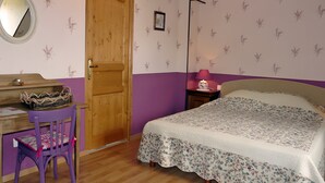 Quarto com porta de madeira, escrivaninha com cadeira, cama com colcha floral e mesa de cabeceira com abajur.
